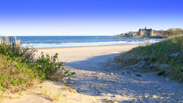 A beach with a sandy path leading to the sea, a wooden fence on the left, and a building with a tower on the right. - Blue Moon Carriage House (Narragansett)