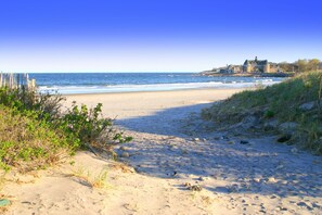Una platja amb un camí de sorra que condueix a la mar, una tanca de fusta a l'esquerra i un edifici amb una torre a la dreta.