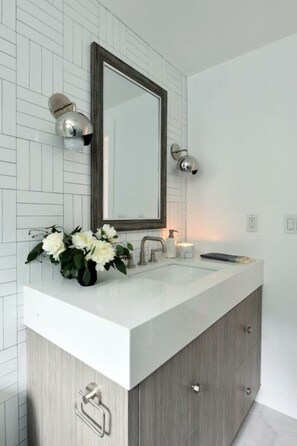 Un baño con salpicadero de azulejos blancos, un tocador de madera con espejo, un lavamanos y un jarrón con flores blancas.
