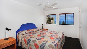 A bedroom with a bed, a ceiling fan, two windows with blinds, a nightstand, and a lamp.