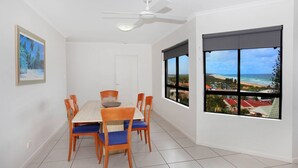 A dining area with a table and chairs, a ceiling fan, and a large window with a view of a beach and ocean.