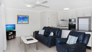 A modern living room with a sofa set, a coffee table, a TV, and a kitchen area with cabinets and appliances.