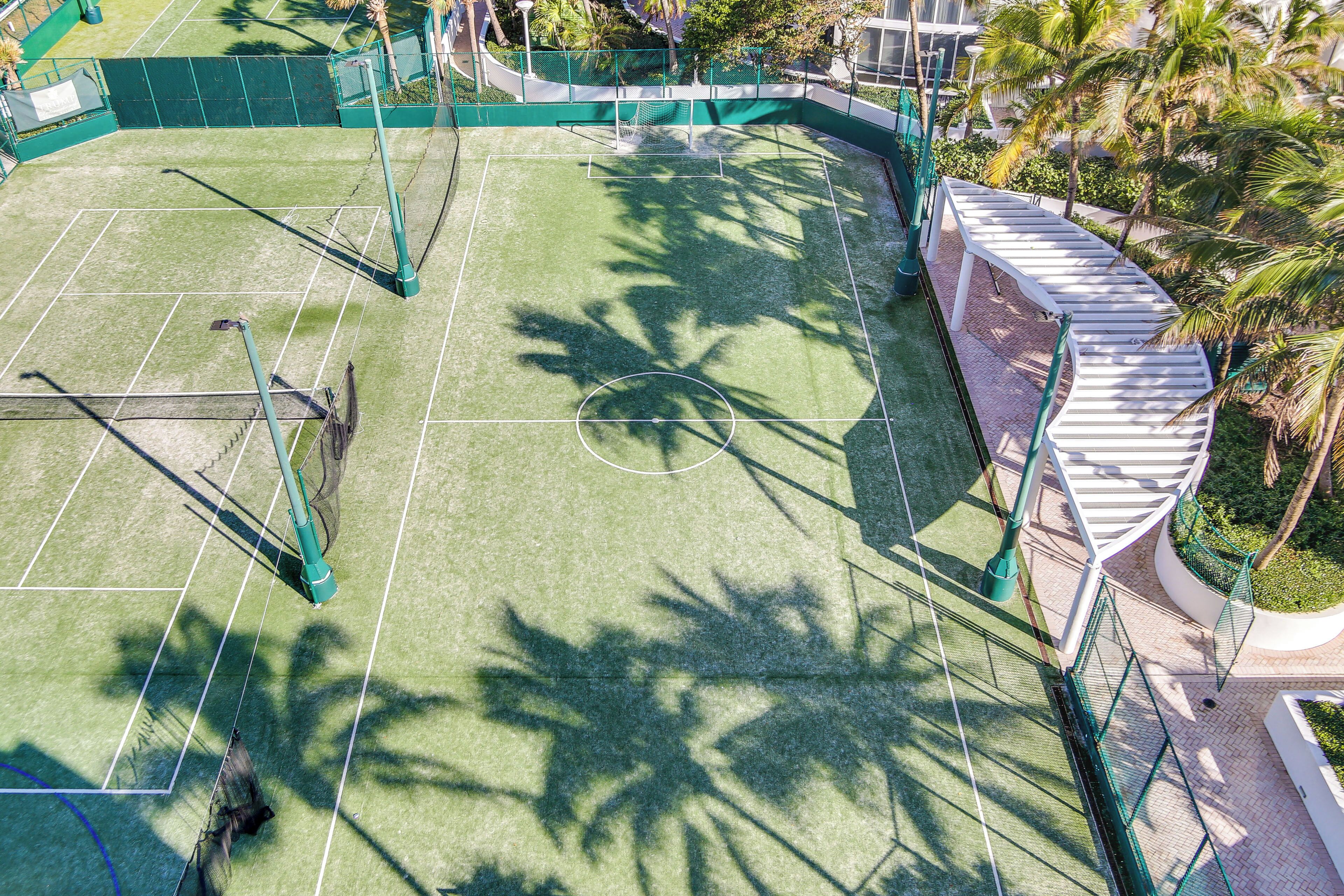 An outdoor tennis court with a net, surrounded by a green fence and palm trees.