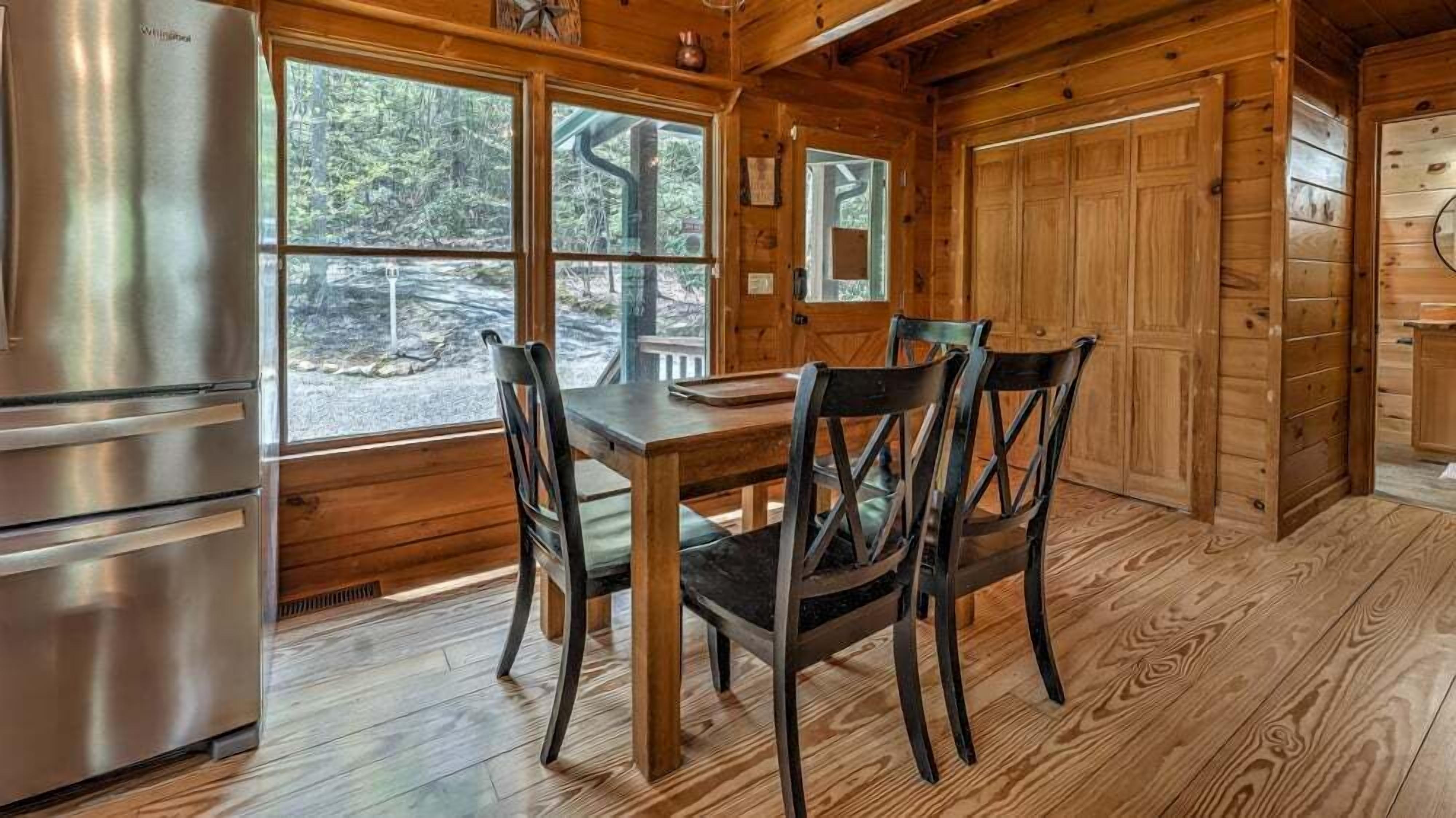 A wooden cabin interior with a dining table, chairs, a refrigerator, and large windows offering a view of the outdoors.