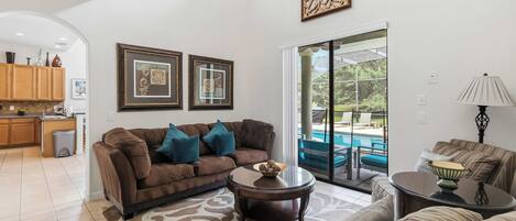 A spacious living room with a brown sofa, a coffee table, and a view of a swimming pool through a glass door.