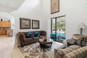A spacious living room with a brown sofa, a coffee table, and a view of a swimming pool through a glass door. - Luxurious 5BR Villa Your Disney Getaway (Kissimmee)
