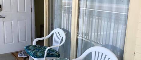 Un balcón con dos sillas de plástico blanco y cojines estampados, una puerta blanca y una ventana con cortinas ligeras.