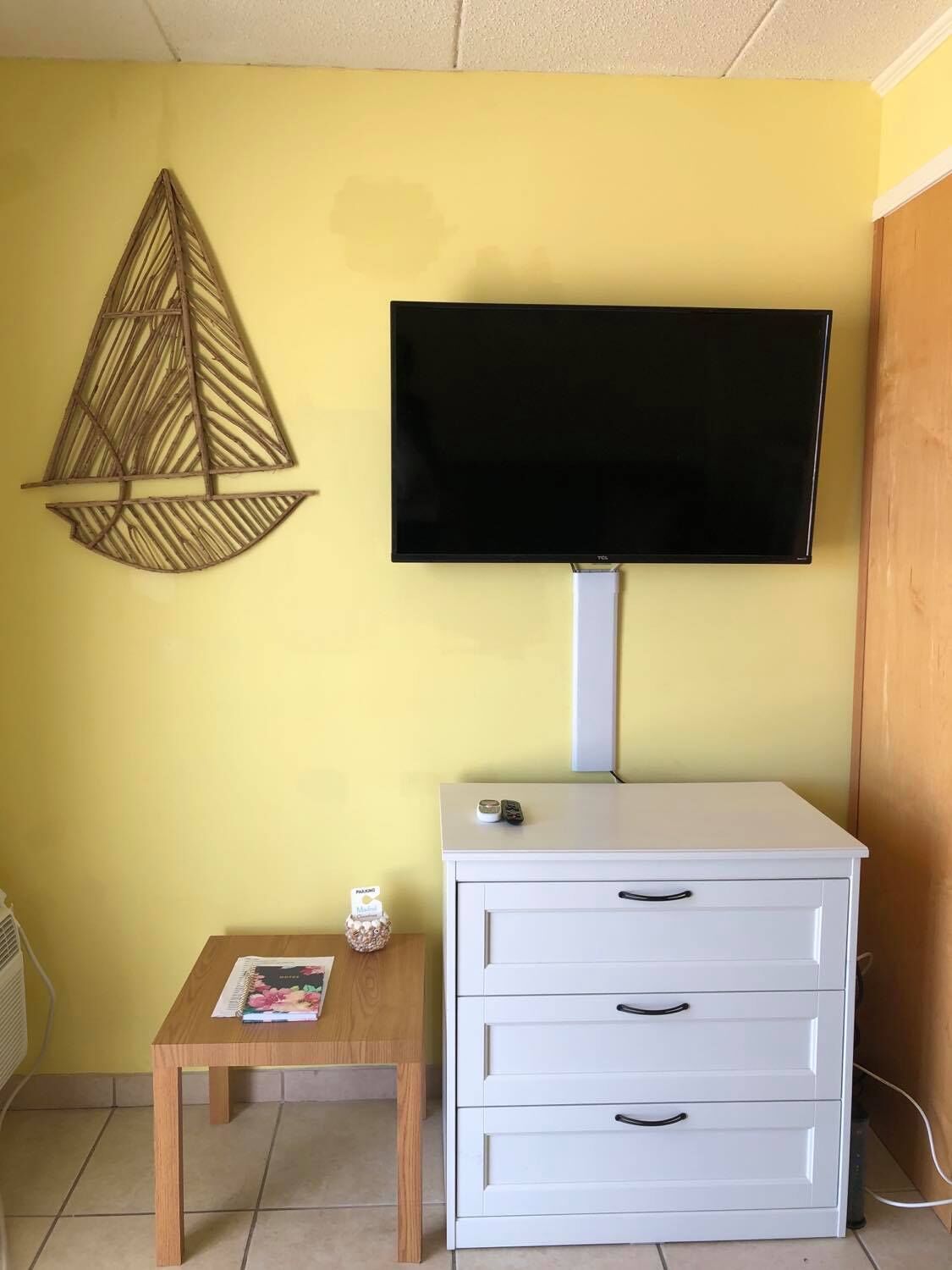 Una habitación con una pared amarilla, un televisor de pantalla plana montado en un mueble blanco, una pequeña mesa de madera con una revista, y un adorno colgando en la pared.