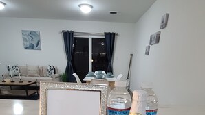 A framed welcome sign, sparkling water bottles, and a bottle of Martini wine on a marble countertop in a well-lit living area with a sofa and dining table in the background.
