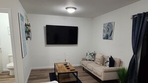 A modern living room with a white sofa, a coffee table, and a dining table set with blue plates and cups. A flat-screen TV is mounted on the wall, and there is a painting above the sofa.