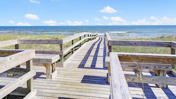 Een houten vlonder die naar een strand leidt, onder een heldere lucht.