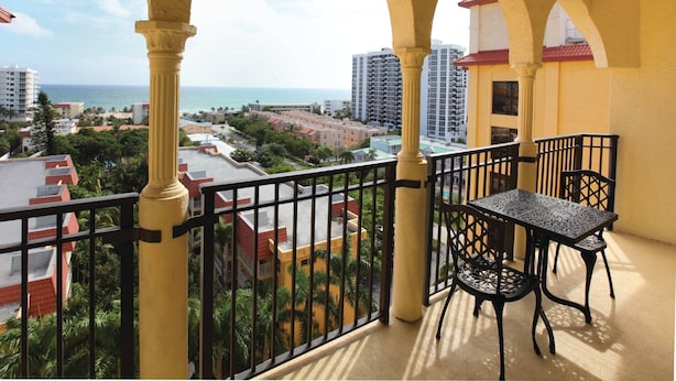 Un balcón con mesa y sillas, con vista a la costa, edificios y cielo despejado.