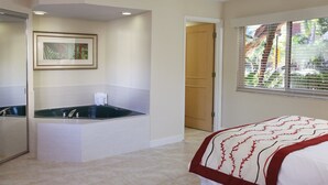 Habitación de hotel con una bañera grande, una cama con colcha de rayas rojas y blancas, un cuadro enmarcado en la pared y una ventana con persianas.