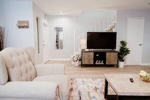 A living room with a sofa, a coffee table, a TV on a wooden stand, a staircase, and a door.