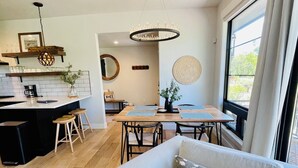 Ein modernes Wohnzimmer mit grauem Sofa, Couchtisch und Essbereich mit Holztisch und Stühlen. Es gibt eine Küchenzeile mit schwarzer Arbeitsplatte und einer Pendelleuchte.
