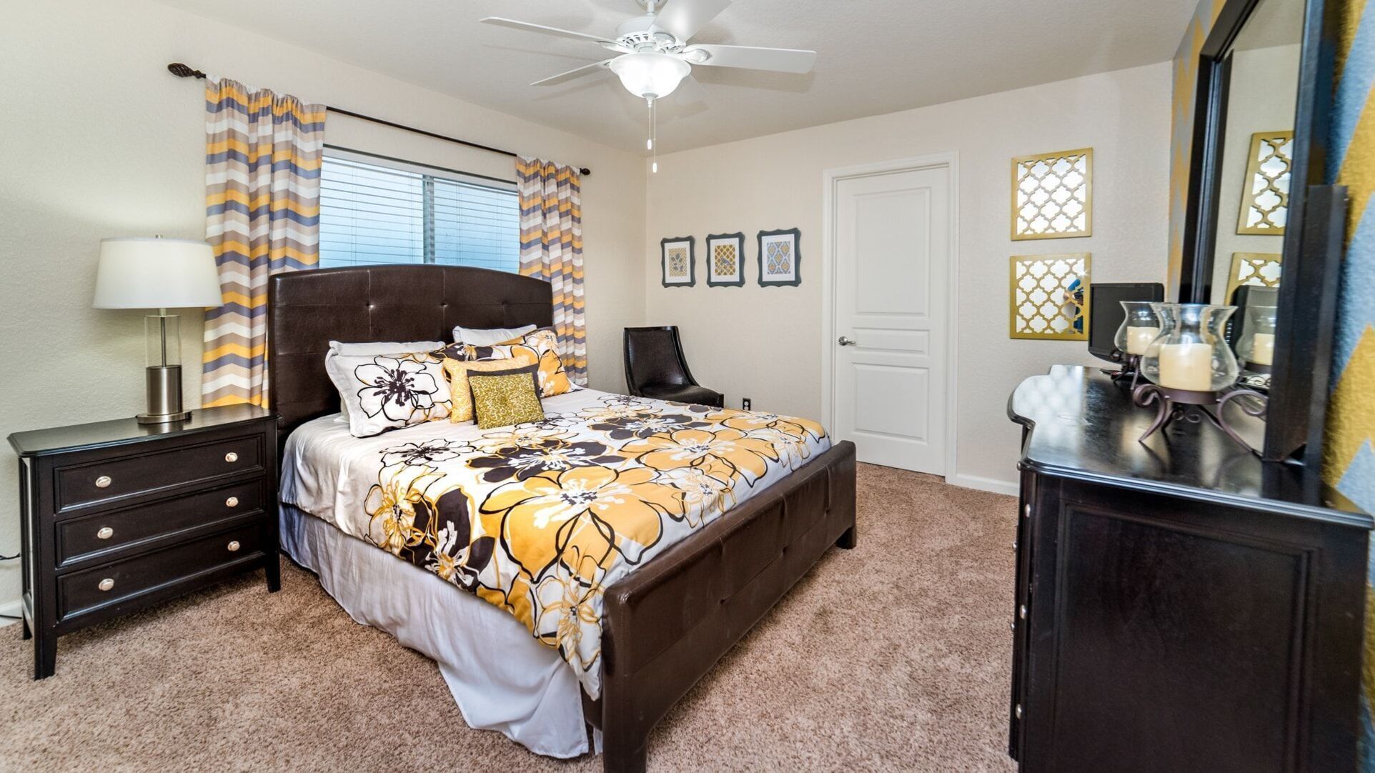 Quarto com cama, penteadeira, mesa de cabeceira, ventilador de teto e janela com cortinas.