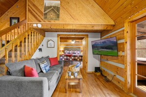 A cozy cabin interior with a wooden staircase, a gray sofa with red and blue cushions, a television mounted on the wall, and a wooden dining table with a basket. - Secluded, Yet Convenient to All Attractions! (Sevierville)