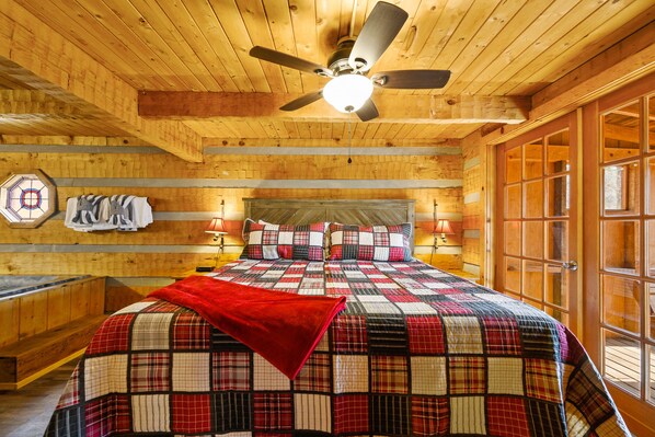A wooden cabin bedroom with a plaid bedspread, a ceiling fan, and a large window. - Secluded, Yet Convenient to All Attractions! (Sevierville)