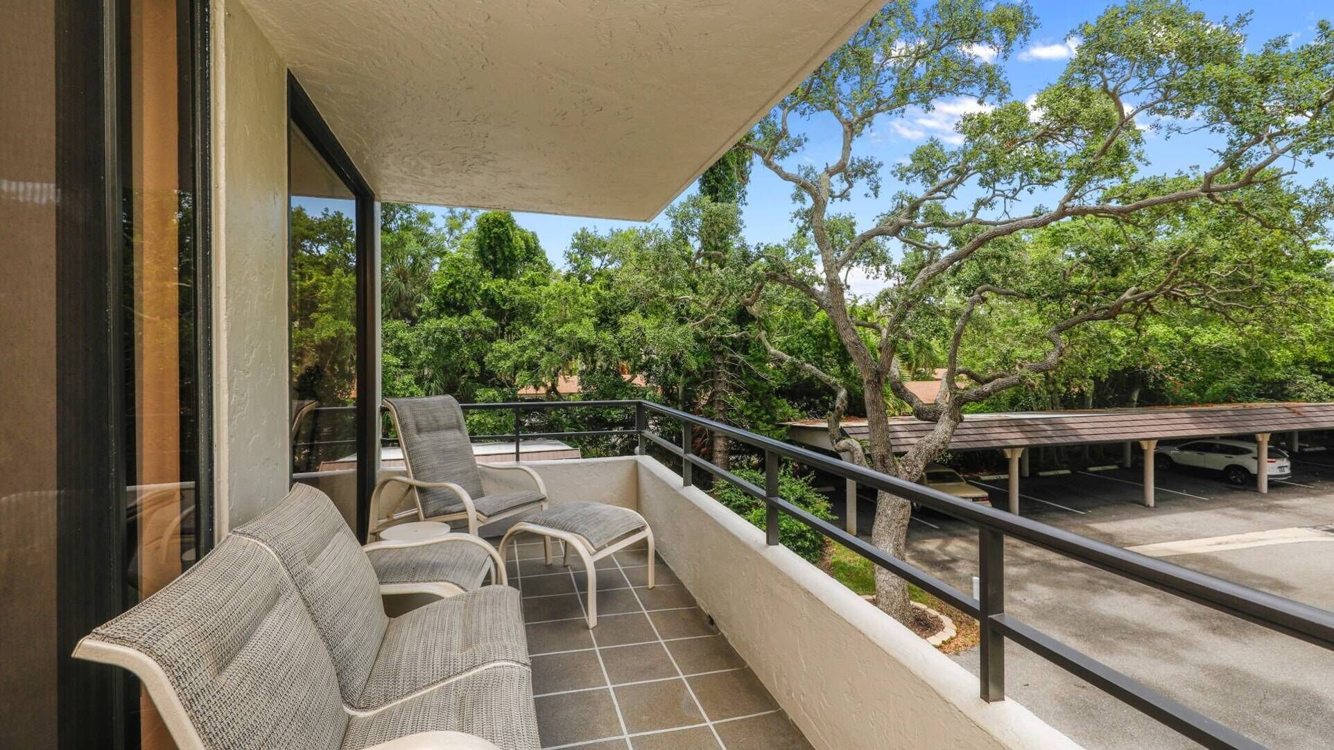 A balcony with two wicker chairs and a small table, overlooking a parking area and lush greenery.