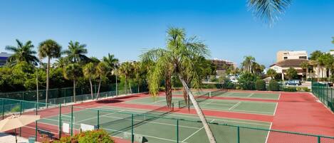 Una cancha de tenis con valla verde, palmeras y un cielo azul despejado.