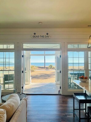 Quarto com vista para a praia, um letreiro com a frase "SEAS THE DAY" e uma área de refeições com mesa e cadeiras.