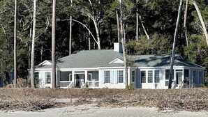 A two-story house with a porch, surrounded by tall trees and a sandy beach.