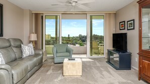A living room with a grey leather sofa, a light blue armchair, a wooden coffee table, a flat-screen TV on a stand, and a balcony with a view of greenery and buildings. - Perfect 2 Bedroom Apartment on The Anchorage, Siesta Key Apartment 1020 (Siesta Key)