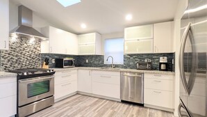A modern kitchen with white cabinets, a stainless steel refrigerator, a microwave, a stove, and a dishwasher. - Cute Edmonds Bowl Cottage (Edmonds)