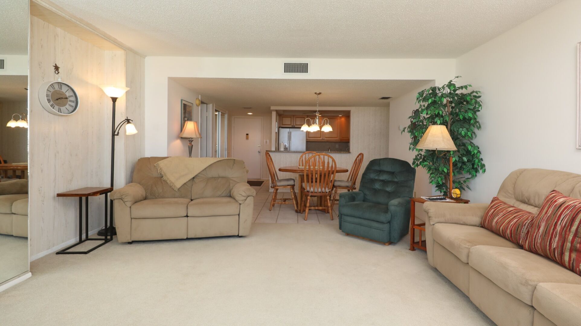 A living room with a beige sofa, a green armchair, a dining table with chairs, and a kitchen in the background.