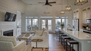 A spacious living area with a fireplace, a sofa, a coffee table, and a TV mounted on the wall. There is a kitchen area with a bar and stools. - Angel Oak Cottage-5bdrm Luxury and Beachfront-4 Pass Golf Cart Included (Daufuskie Island)