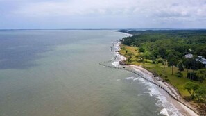 Vista aérea de una zona costera con una playa larga y estrecha, un muelle de madera y un área boscosa con edificios.