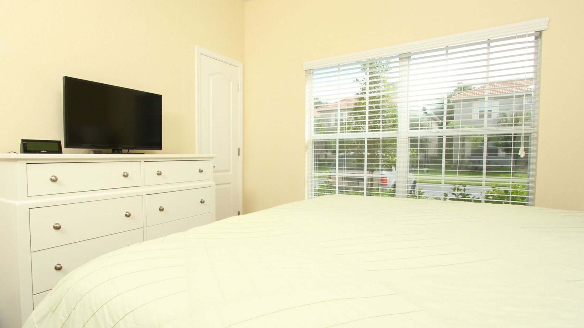 A bedroom with a bed, a dresser with drawers, a television on top, and a window with blinds.