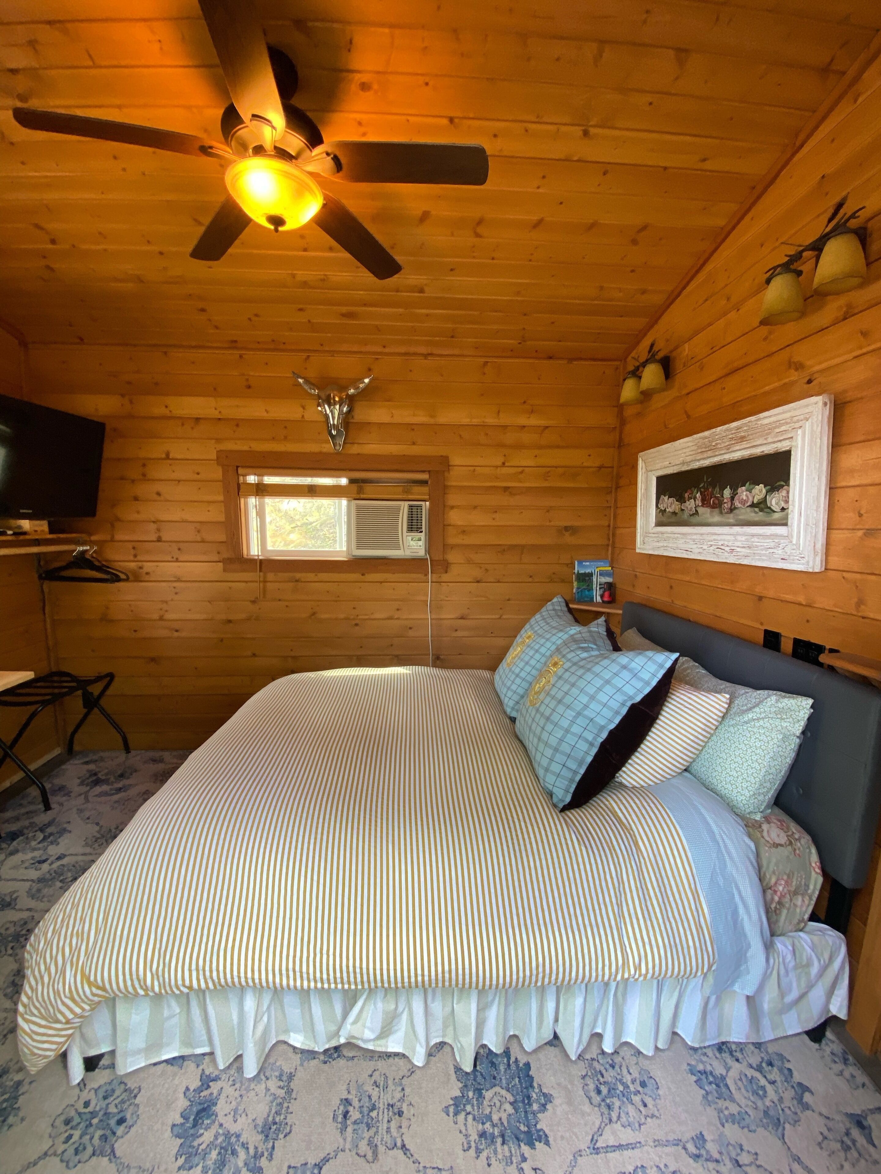 A wooden cabin bedroom with a bed, a ceiling fan, a mounted TV, and a window with an air conditioner.