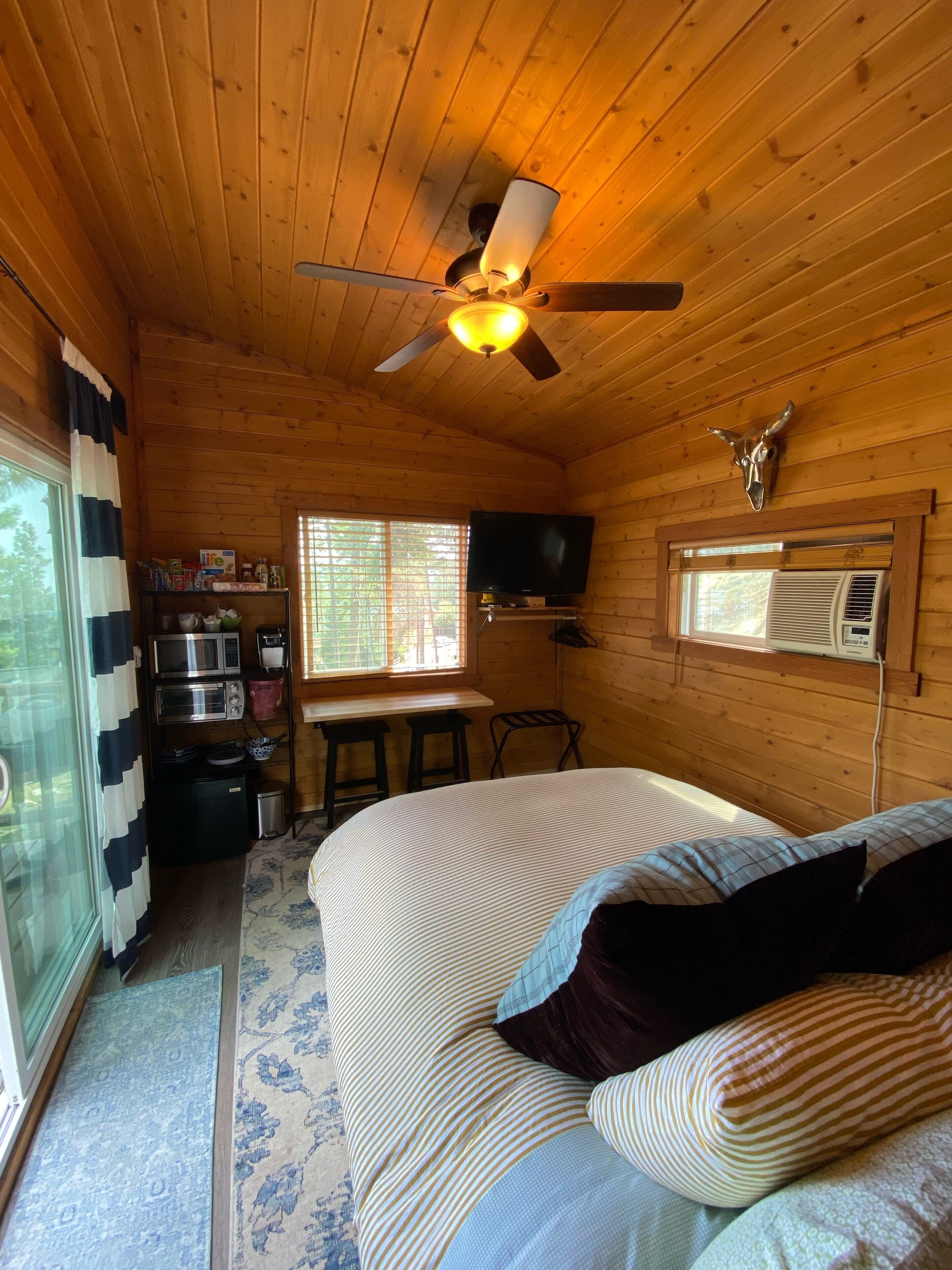 Una habitación tipo cabaña acogedora, con una cama, un ventilador de techo, un televisor montado y una ventana con persianas.