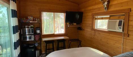 Una habitación tipo cabaña acogedora, con una cama, un ventilador de techo, un televisor montado y una ventana con persianas.