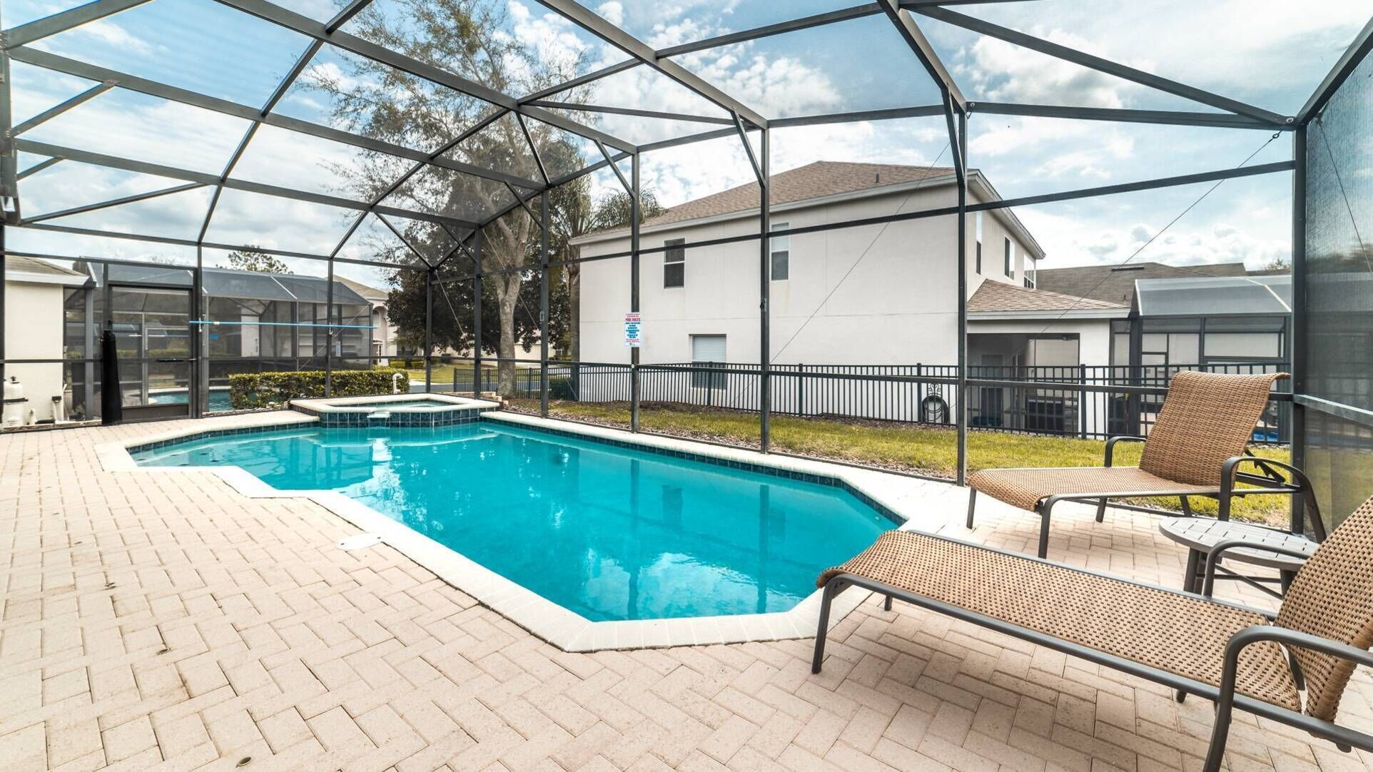 A covered outdoor swimming pool with a brick patio, surrounded by a metal fence and a building in the background.