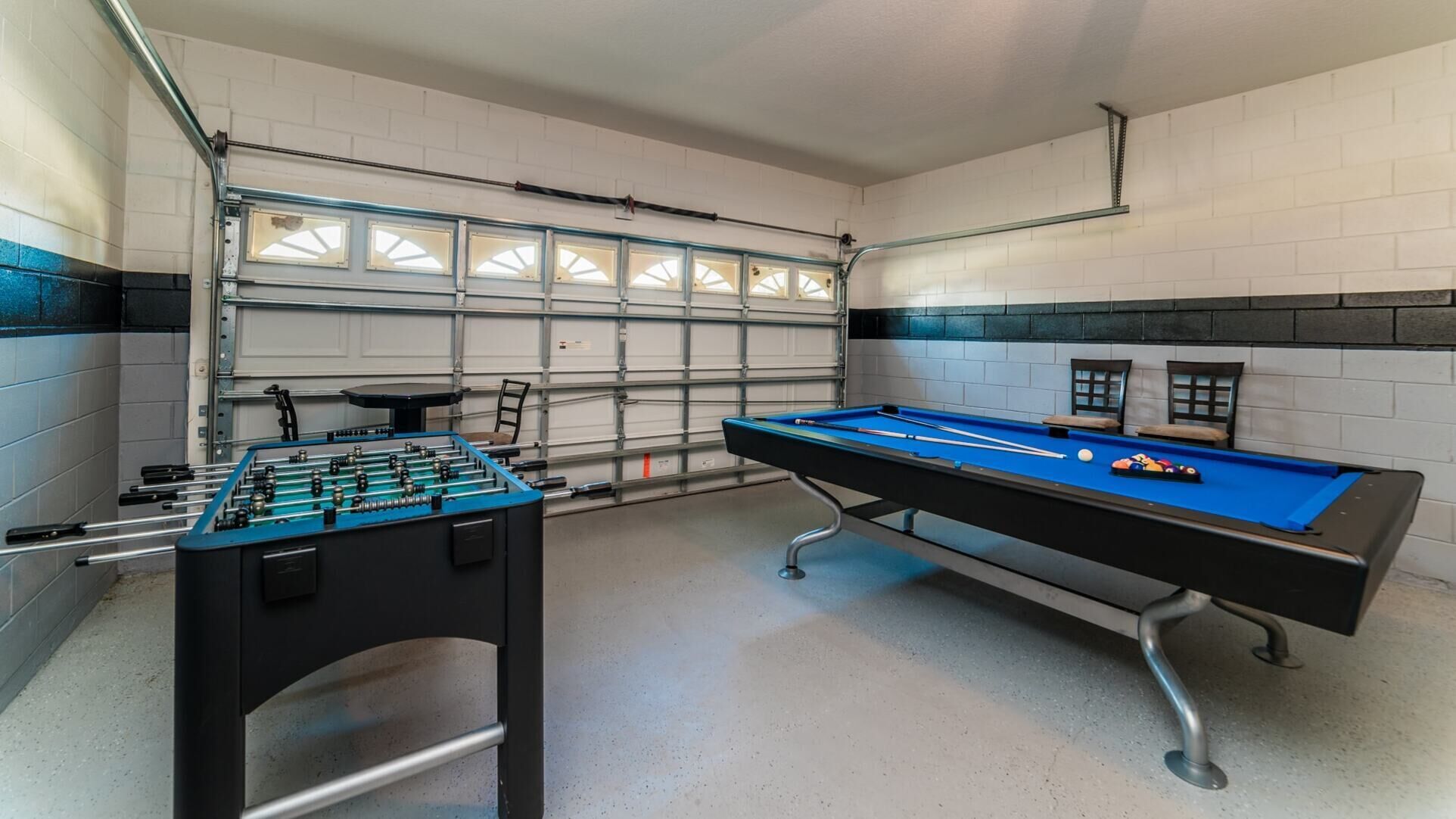 A pool table with foosball and pool cues, a table tennis table, and chairs in a room with white tiled walls and a garage door in the background.