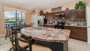 A kitchen with a granite island, dark wooden cabinets, stainless steel appliances, and a dining area with a glass table and chairs. - Exclusive 5 Star Villa with Private Pool on Windsor Palms Resort, Orlando Villa 4882 (Orlando)