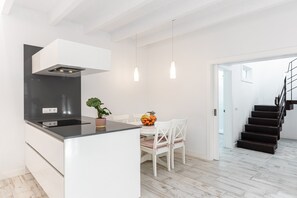 A modern kitchen with a central island, white cabinetry, a black range hood, pendant lights, a dining table with chairs, and a staircase. - Holiday Home ‘Es Raco’ with Mountain View, Jacuzzi, Wi-Fi, Air Conditioning & Terraces (Costitx)
