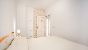 Een hotelkamer met een bed, een nachtkastje met een lamp, een deur met decoratief paneel, en uitzicht op de gang.