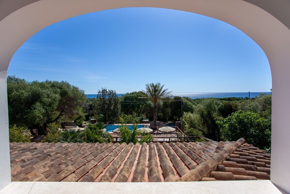A view through an archway of a rooftop with a swimming pool, surrounded by greenery and a clear sky.