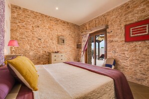 A bedroom with a stone wall, a bed with a white bedspread, a nightstand with a lamp, and a view through a glass door.