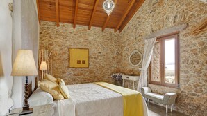 A bedroom with stone walls, a wooden ceiling, a bed with a yellow blanket, a bedside table with a lamp, and a window with curtains.