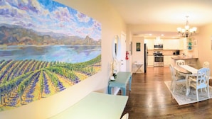 A dining area with a table and chairs, a kitchen in the background, and a large landscape painting on the wall. - Spacious Westside  Wine Trail Retreat ★★★★★ (West Kelowna)