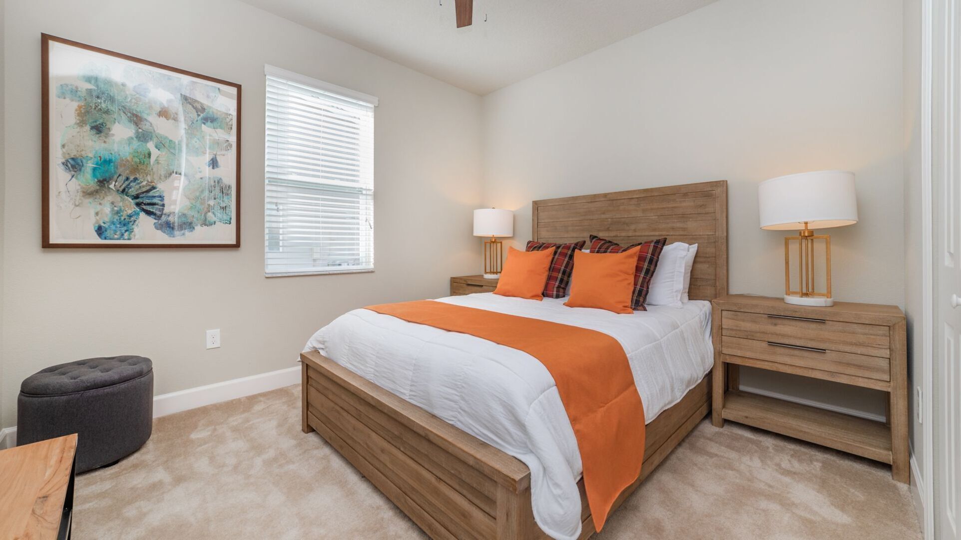 A bedroom with a wooden bed, two bedside tables, a nightstand, a grey ottoman, and a large abstract painting on the wall.