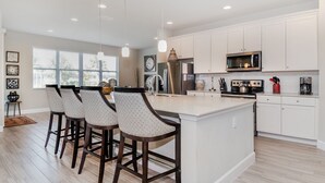 Una cocina moderna con armarios blancos, una isla central con taburetes altos, electrodomésticos de acero inoxidable e iluminación colgante.