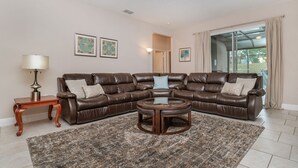 A living room with a brown leather sectional sofa, a small wooden side table, a round coffee table, and a patterned rug. There are two framed pictures on the wall and a window with curtains.