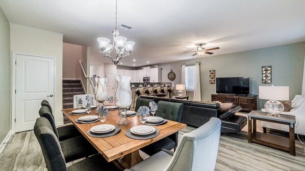 A dining area with a wooden table set for six, a chandelier, and a kitchen in the background.