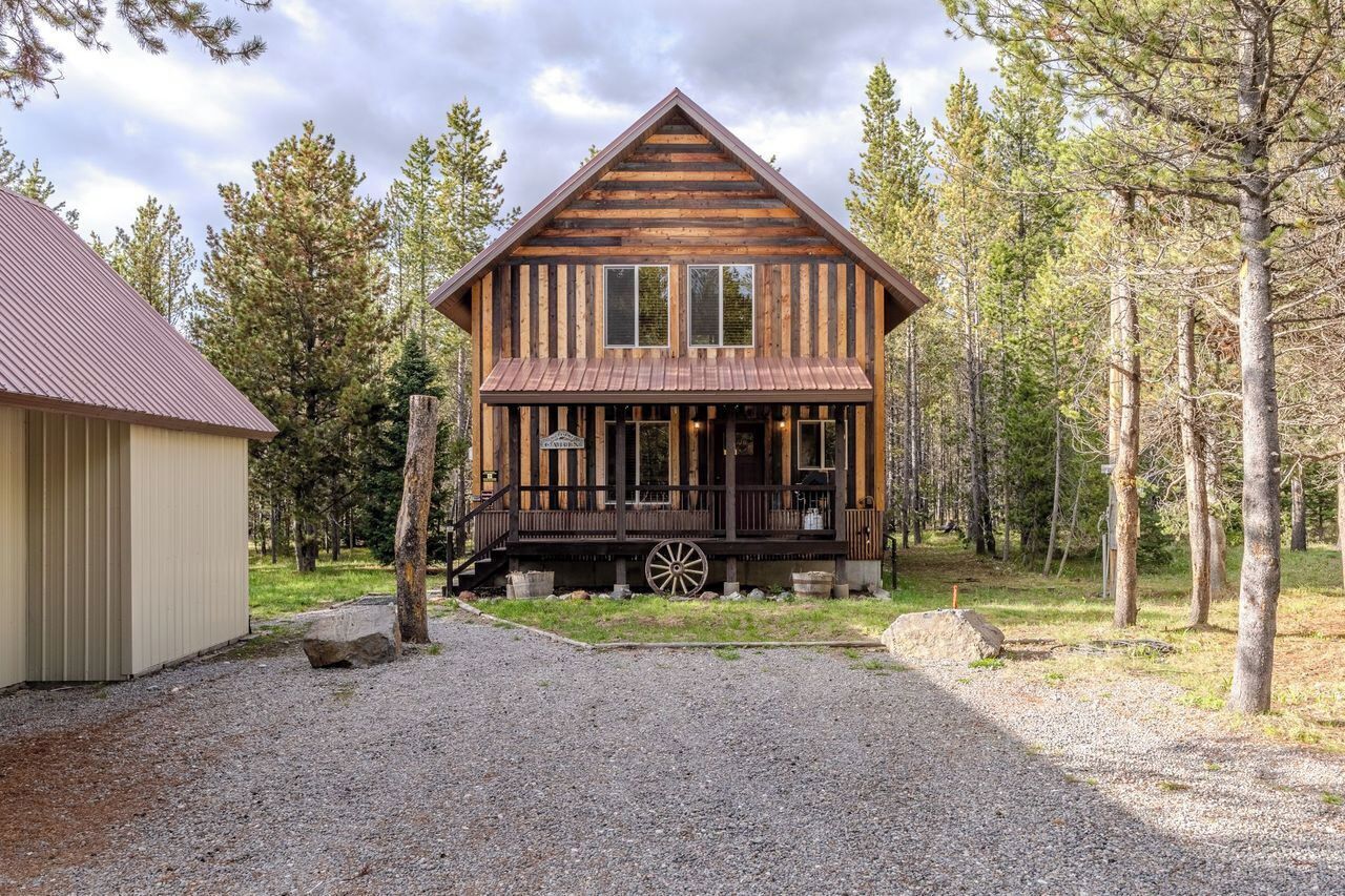 Une cabane rustique avec une façade en bois, une véranda ornée d’une roue de charrette et une allée gravillonnée.