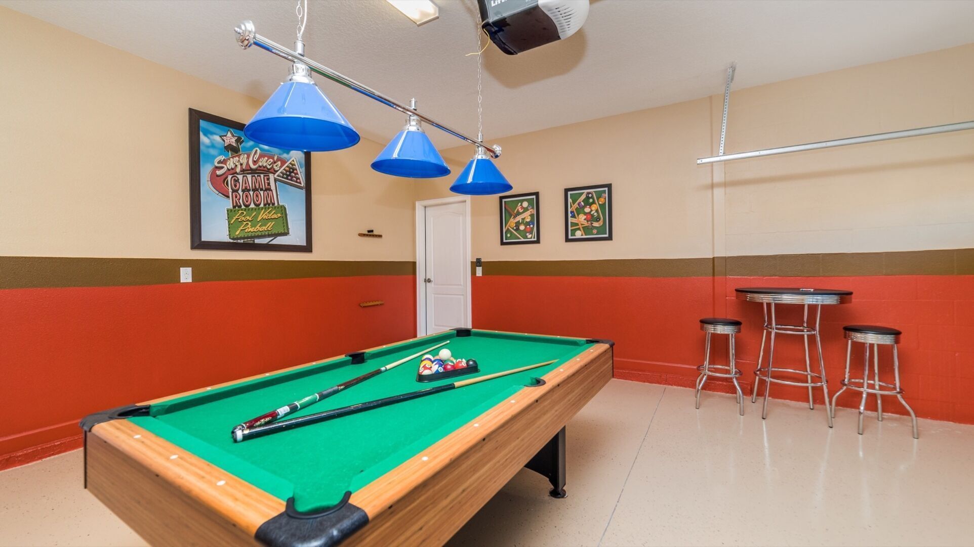 Une table de billard recouverte d’un drap vert, entourée de murs rouges et d’une porte blanche. Trois lampes bleues sont suspendues au-dessus de la table, et une affiche encadrée est accrochée au mur, à gauche.